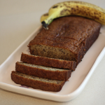 Image of freshly baked banana bread partially sliced on a platter with a banana in the background